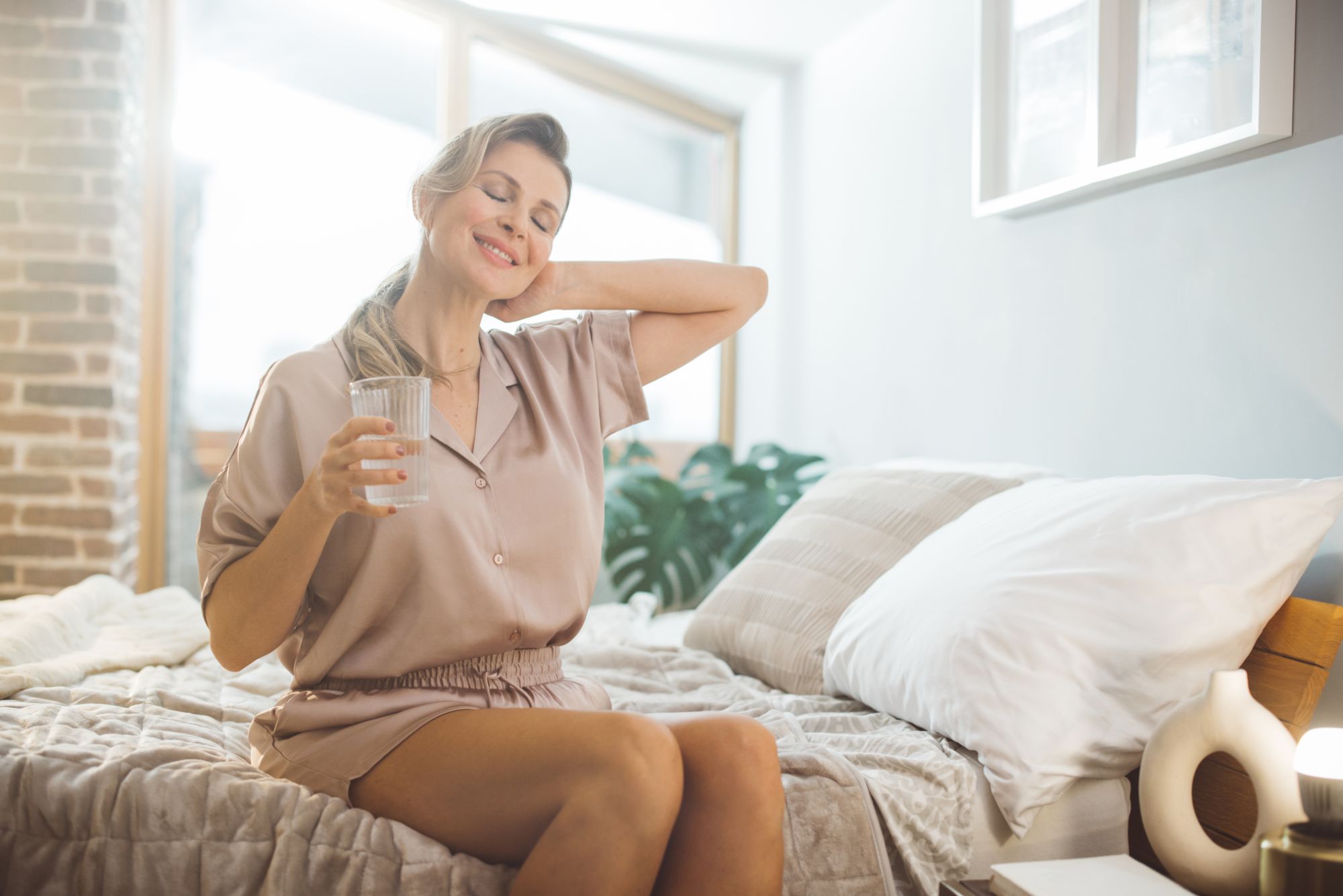 A woman experiences the balanced start provided by hormone replacement therapy, sitting on her bed in the morning drinking a glass of water
