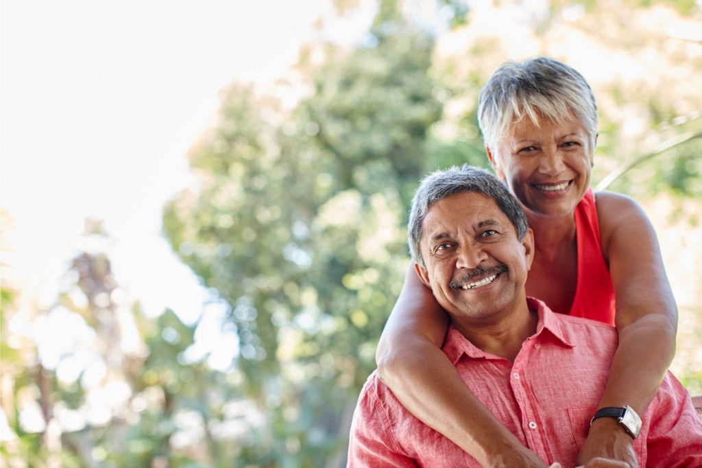 A senior man and woman smiling at the camera with the woman wrapping her arms around the man's shoulders