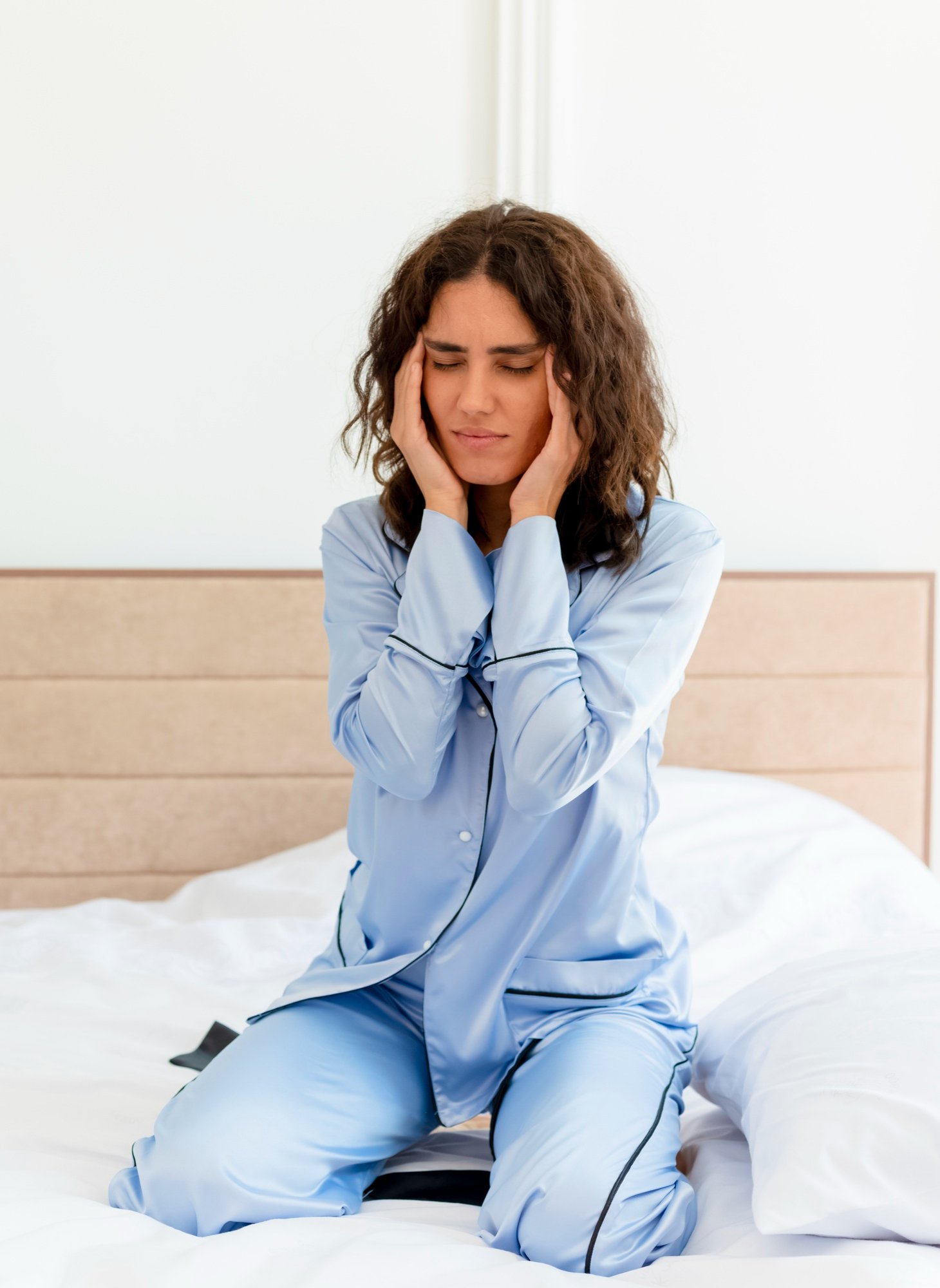 Woman in blue pajamas sitting on bed touching her temples while experiencing headaches and fatigue related to HPA axis dysfunction.